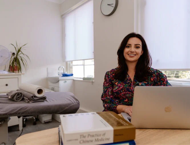 Katie Siggins is an Acupuncturist based in Clapham near Balham, Battersea & Wandsworth, South West London. She is pictured sitting at a desk with her laptop.